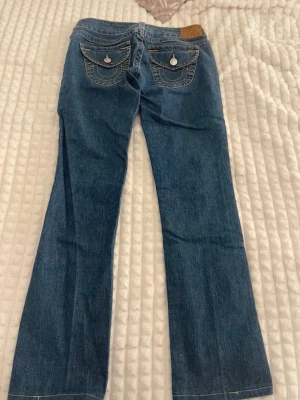 Blå lowwaist jeans från True Religion - Säljer ett par klassiska blå jeans från True Religion med ikoniska kontrastsömmar och snygga ficklock bak med vita knappar. Jeansen har låg midja och är tillverkade i slitstark denim. Perfekta för dig som gillar en avslappnad men trendig look.