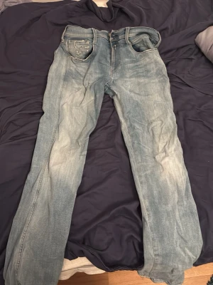 Ljusblå raka jeans med slitningar - Säljer ett par ljusblå jeans med raka ben och klassisk femficksdesign. Jeansen har en snygg tvättad look med slitningar och ljusare partier längs benen. Perfekta för en avslappnad stil. Materialet är denim i bomull.