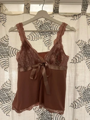 Rosa spetslinne med satinrosett - Sött pyjamaslinne i dammig rosa med spetsdetaljer på axelband och byst. Satinband under bysten som knyts till rosett framtill. Mjuk och lätt materialblandning, perfekt för mysiga kvällar. Spetskant nertill för extra romantisk känsla.