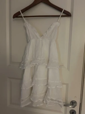 Vit volangtop från Gina Tricot - Säljer en vit linnetopp från Gina Tricot med smala axelband och flera lager volanger. Toppen har spetsdetaljer och en v-ringad hals. Perfekt för varma dagar och har en luftig känsla tack vare det tunna materialet. Kan även användas som kort klänning. (Säljer då den aldrig andvänds)