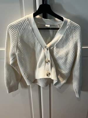 Vit kofta från Lindex - Snygg vit stickad kofta från Lindex med tre stora pärlemorknappar framtill. Koftan har ribbstickade detaljer, v-ringad hals och långa ärmar. Perfekt för lager på lager och enkel att styla till olika outfits.