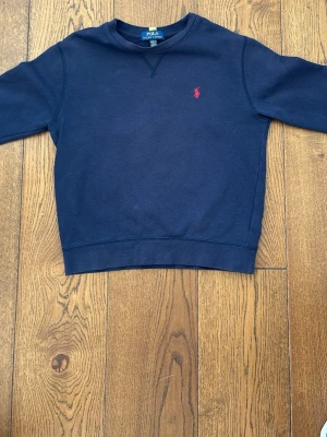 Mörkblå sweatshirt från Polo Ralph Lauren - Mörkblå sweatshirt från Polo Ralph Lauren med klassisk rund halsringning och röd broderad logga på bröstet. Tröjan har ribbade muddar och är tillverkad i mjukt bomullsmaterial. Perfekt för en avslappnad och stilren look. Hör av er vid frågor, pris kan diskuteras 