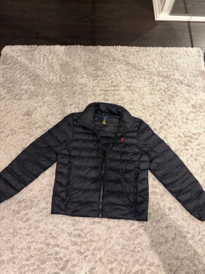 Svart pufferjacka från Polo Ralph Lauren - Snygg svart pufferjacka från Polo Ralph Lauren med klassisk röd logga på bröstet. Jackan har dragkedja framtill, två sidofickor med dragkedja och hög krage. Perfekt för kalla dagar och enkel att matcha med olika outfits. Köpt för en månad sedan. Storlek XL i barnstorlekar så storlek S i herr!