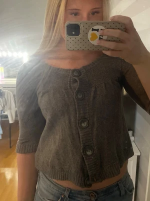 Kort grå kofta med stora knappar - Säljer en kort grå kofta med trekvartsärmar och stora dekorativa knappar framtill. Koftan har ribbade muddar och en rund halsringning. Perfekt att slänga över en topp för en chill look.