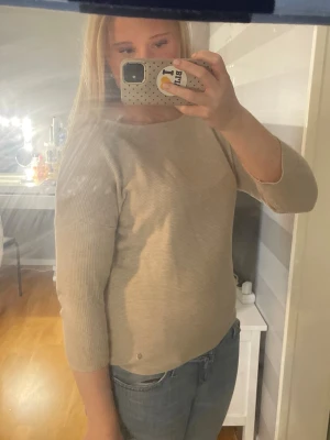 Beige ribbstickad tröja med 3/4-ärm - Säljer en stilren beige ribbstickad tröja med 3/4-ärmar och rund halsringning. Tröjan har en diskret metallknapp vid nederkanten och är gjord i ett mjukt material som passar perfekt till jeans.