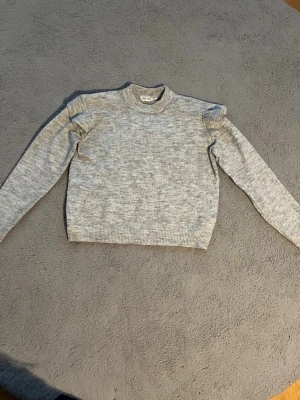 Grå stickad tröja med volangdetalj - Snygg gråmelerad stickad tröja med volangdetaljer vid axlarna. Tröjan har rund halsringning, långa ärmar och ribbstickade muddar vid ärmslut och nederkant. Lite nopprig. Mått: Längd 50cm, Bredd under armarna 50cm, Bredd nertill 41cm. Material: 52% Polyester, 41% Akryl, 4% Ull, 3% Elastan.