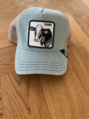 Ljusblå keps från The Farm med ko - Snygg ljusblå keps från The Farm med beige mesh baktill och broderad patch framtill med en ko och texten 'CASH’. Den är justerbar så passar alla. 