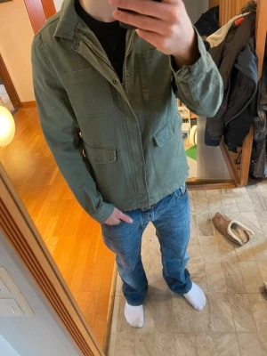 Fieldjacket/overshirt/utilityjacket - Snygg olivgrön utilityjacka med fyra framfickor och klassisk krage. Jackan har rak passform och stängs med knappar framtill. Perfekt för lager-på-lager och enkel att styla till jeans eller chinos. OBS tvättlappen är avklippt! Den är i jeans material.