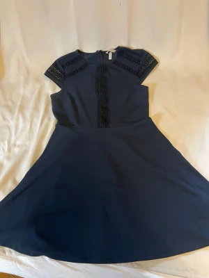 Marinblå kortklänning med spetsdetaljer - Snygg marinblå kortklänning med spetsdetaljer på axlar och längs bröstet. Klänningen har korta ärmar och en rund halsringning. Perfekt för dig som gillar stilrena och eleganta plagg.