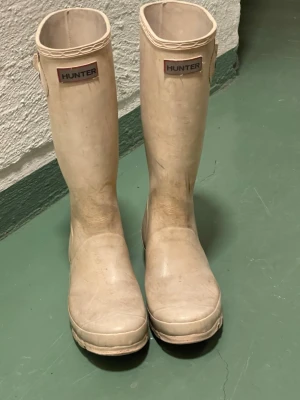 Vita höga gummistövlar Hunter storlek 38.5 - Stilrena vita gummistövlar från Hunter med klassisk hög modell och justerbart spänne vid skaftet. Perfekta för regniga dagar och festivaler. Hunter-logga framtill och grov sula för bra grepp. Storlek 38.5