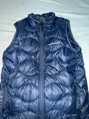 Mörkblå dunväst Peak Performance - Snygg mörkblå dunväst från Peak Performance med hög krage och dragkedja framtill. Västen har två sidofickor med dragkedja och quiltad design som ger en sportig look. Perfekt för lager-på-lager och håller dig varm under kyliga dagar.