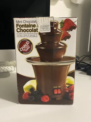 Mini chokladfontän röd - Säljer en mini chokladfontän i röd och svart plast med metallkant. Fontänen har två knappar för motor och värme samt en EU-kontakt. Perfekt för att doppa frukt eller godis i smält choklad. Originalkartong medföljer.