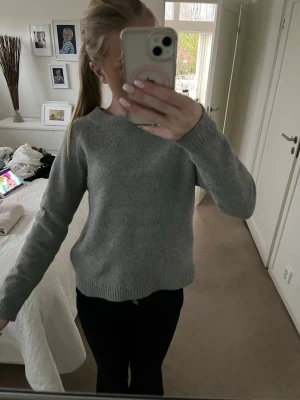 Grå stickad tröja med rund hals - En enkel och stilren grå stickad tröja med rund hals och ribbade muddar vid ärmslut och nederkant. Perfekt att matcha med jeans eller leggings. Materialet ser ut att vara mjukt och bekvämt, och tröjan har en klassisk passform.