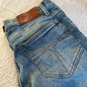 Blå jeans från Tiger of Sweden - Säljer ett par klassiska blå jeans från Tiger of Sweden i modellen Evolve. Jeansen har raka ben, snygga slitningar och kontrastsömmar. De är tillverkade i bomull och har en läderpatch bak i midjan. Perfekta för dig som gillar stilrena och tidlösa jeans.