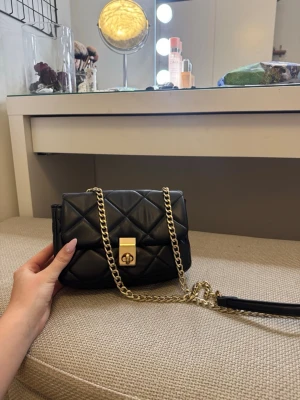 Svart quiltad handväska med guldkedja - Snygg svart quiltad handväska med guldfärgad kedjerem och vridlås framtill. Väskan har en klassisk rektangulär form och är tillverkad i konstläder. Insidan har ett fack med dragkedja för småsaker. Perfekt accessoar för att lyfta din outfit.