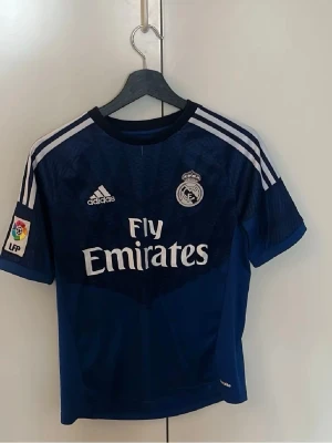 Real Madrid målvaktströja Navas #13 - Mörkblå Real Madrid målvaktströja från Adidas med vita detaljer och tryck. Tröjan har klubbmärke, Adidas-logga, Fly Emirates sponsortryck och LFP-märke på ärmen. På ryggen står det Navas och nummer 13. Tillverkad i funktionsmaterial.