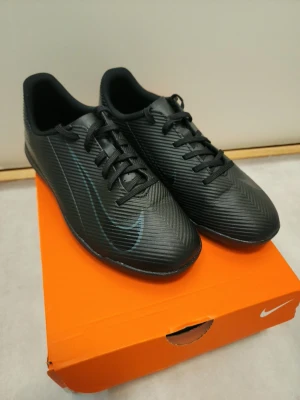 Svarta Nike fotbollsskor med snörning - Snygga svarta fotbollsskor från Nike med diskret mönster och klassisk snörning. Skorna har en låg profil och är tillverkade i syntetmaterial med en stilren design och subtil Nike-logga på sidan. Perfekta för dig som vill ha en clean look på planen.