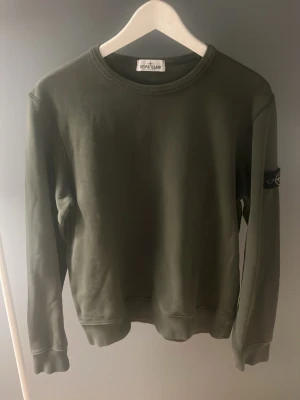 Olivgrön sweatshirt från Stone Island - Säljer en olivgrön sweatshirt från Stone Island med klassisk rund hals och ribbade muddar. Tröjan har den ikoniska Stone Island-loggan på vänster ärm. Perfekt för dig som gillar streetwear och stilrena plagg.