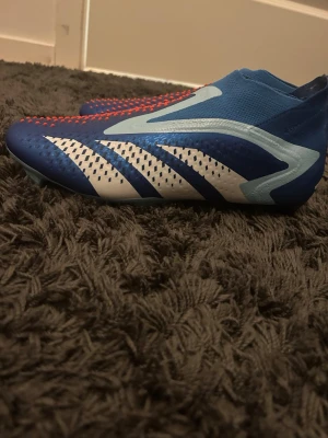 Blåa Adidas Predator fotbollsskor Elite med påse och låda - Säljer ett par blåa Adidas Predator fotbollsskor med vita och ljusblå detaljer samt röda prickar på ovandelen. De är helt nya med både påse och låda Skorna har en strumpliknande passform utan snörning och är tillverkade i syntetmaterial för bra grepp och kontroll på planen. 