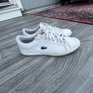 Vita sneakers från Lacoste - Säljer ett par klassiska vita sneakers från Lacoste med snörning och den ikoniska krokodilloggan på sidan. Skorna är i skinn och har en stilren design med rund tå och platt sula. Perfekta för en clean och sportig look.