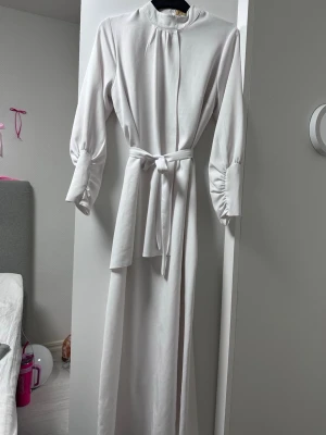 Vit långklänning med knytband - Elegant vit långklänning med höghalsad krage, långa ärmar och knytband i midjan. Klänningen har diskreta veck vid axlarna och breda manschetter med knappar. Perfekt för dig som gillar stilrena och klassiska plagg.