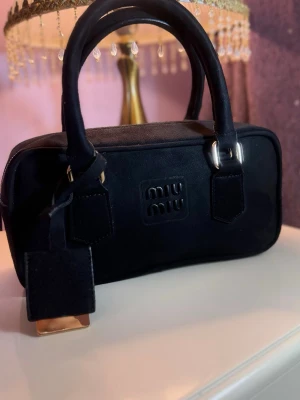 Svart handväska från Miu Miu - Snygg svart handväska från Miu Miu med rektangulär form och dubbla handtag. Väskan har en diskret logotyp på framsidan och detaljer i silverfärgad metall. Tillverkad i ett tygmaterial som ger en stilren och modern look. Perfekt accessoar för att lyfta din outfit.