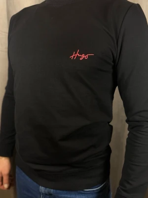 Svart långärmad topp från Hugo - Stilren svart långärmad topp från Hugo med röd broderad logga på bröstet. Klassisk rund halsringning och normal passform. Perfekt till jeans eller under en jacka för en clean look.