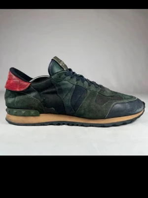 Valentino rockrunners - Snygga sneakers från Valentino Garavani i mörkgrön mocka och svart textil med röda detaljer på hälen. Skorna har svarta snören, beige sula och coola nitar på baksidan. Perfekt för dig som gillar exklusiva och unika sneakers.