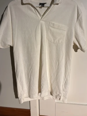 Vit frottéskjorta från H&M - Vit skjorta från H&M i mjuk frotté med kort ärm och bröstficka. Regular fit med klassisk krage och enkel, clean look. Perfekt för dig som gillar avslappnad stil och vill ha något bekvämt och svalt.