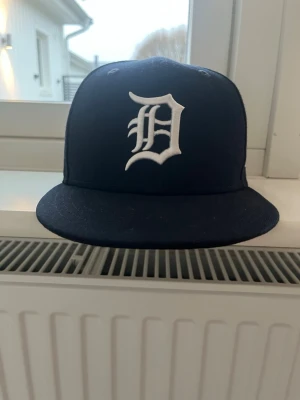 Svart keps med vit D-logga - Snygg svart keps med platt skärm och en stor vit broderad D-logga framtill. Klassisk streetwear-stil, perfekt för att lyfta din outfit. Justerbar baktill för bästa passform. Materialet är troligtvis bomull eller polyester.