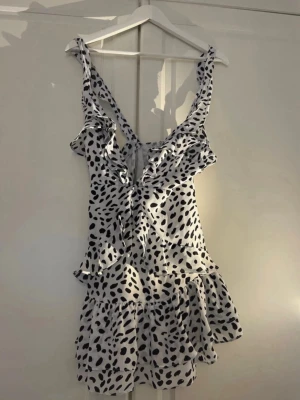  prickig volangklänning - Söt kort klänning i vitt med svarta prickar och volangdetaljer. Klänningen är ärmlös med breda axelband och har en lätt v-ringning framtill. Perfekt för varma dagar och har volanger nertill.