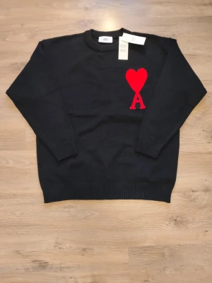 Svart stickad tröja från Ami Paris - Svart stickad tröja från Ami Paris med rund halsringning och ribbade muddar. På bröstet finns ett stort rött hjärta med bokstaven A. Tröjan har en klassisk passform och är tillverkad i mjukt material. Perfekt för dig som gillar stilrena detaljer.