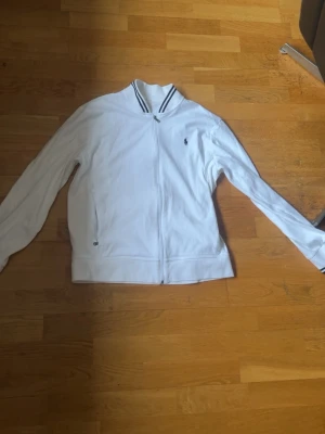 Vit zip hoodie från Ralph Lauren - Snygg vit hoodie från Ralph Lauren med dragkedja framtill och klassisk logga broderad på bröstet. Tröjan har ribbade muddar, två fickor och en krage. Perfekt för en clean och sportig look.