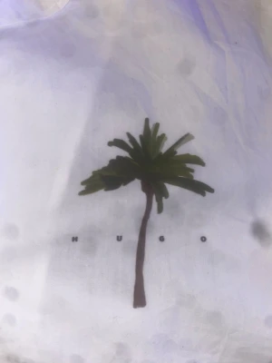 Vit skjorta med palmtryck från Hugo - Säljer en vit skjorta från Hugo med ett stilrent palmtryck på bröstet och klassisk krage. Skjortan har en clean look och är perfekt för dig som gillar minimalistisk design med en twist. Märket syns tydligt under trycket.