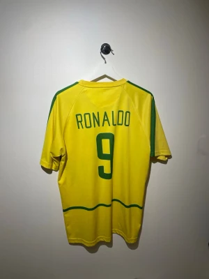 Brasilien Ronaldo 9 fotbollströja Nike - Gul Brasilien fotbollströja från Nike med gröna detaljer, nummer 9 och Ronaldo på ryggen. Klassisk landslagströja med broderad Brasilien-logga och Nike Swoosh på bröstet. Perfekt för dig som älskar fotboll och vill hylla en ikon.