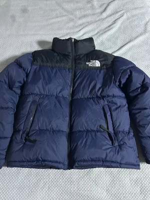 Blå dunväst från The North Face - Snygg blå dunväst från The North Face med svarta axelpartier och hög krage. Västen har dragkedja framtill, två snedställda fickor med dragkedja och logga på bröstet. Perfekt för lager-på-lager och streetwear-stil.