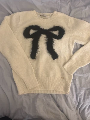 Beige stickad tröja med rund hals - En enkel och stilren vitbeige stickad tröja med rund hals och långa ärmar. Tröjan har ribbstickade muddar vid ärmslut och nederkant. Den har även en fluffig svart rosett i mitten. Perfekt för lager-på-lager och passar till många olika outfits. Den är ifrån Lindex Holly & whyte’s collection. Orginal pris 399kr