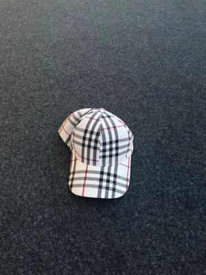Burberry keps - Snygg keps med rutigt mönster i vitt, svart och rött. Kepsen har böjd skärm och justerbar rem bak. Tillverkad i ett mjukt tygmaterial och har en tidlös design som passar till många olika stilar.