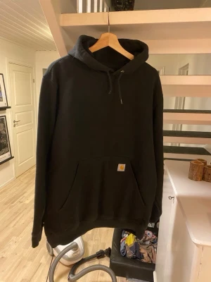 Svart hoodie från Carhartt - Säljer en svart hoodie från Carhartt med klassisk känguruficka och liten Carhartt-logga på fickan. Hoodien har dragsko i huvan och ribbade muddar vid ärmslut och nederkant. Perfekt för en avslappnad och stilren look.