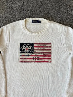 Vit stickad flaggtröja Polo Ralph Lauren - As fet och ovanlig vit stickad vintage tröja från Polo Ralph Lauren med amerikansk flagga och textapplikation framtill. Rund halsringning och ribbade muddar vid ärmslut och nederkant. Perfekt för dig som gillar klassisk och avslappnad stil.