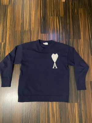 Mörkblå stickad tröja från Ami Paris - Mörkblå stickad tröja från Ami Alexandre Mattiussi med vit hjärtlogga och bokstaven A på bröstet. Tröjan har rund halsringning, ribbade muddar och nederkant. Perfekt för dig som gillar stilrena och trendiga plagg. Ami Paris