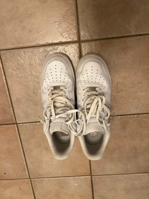 Vita Nike Air Force 1 sneakers - Klassiska helvita Nike Air Force 1 sneakers med lågt skaft. Skorna har perforerad tå för ventilation, kraftig sula och ikonisk Swoosh-logga på sidan. Tillverkade i läder med textilfoder och snörning framtill. Perfekta för en clean och stilren look.