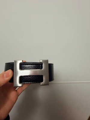 Svart skärp med H-spänne i silver - Stilrent svart skärp med klassiskt H-format spänne i silverfärgad metall. Skärpet är gjort i skinn med vit kontrastsöm längs kanterna, vilket ger en exklusiv känsla. Perfekt accessoar för att lyfta din outfit.