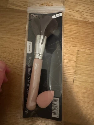 Cien puderborste & makeupsvamp set - Säljer ett 2-pack från Cien med en puderborste och en mini makeupsvamp. Borsten har ett ljusrosa handtag, silverfärgat fäste och mjuka, mörkbruna borst. Makeupsvampen är ljusrosa och droppformad. Perfekt för att applicera puder och blenda foundation.