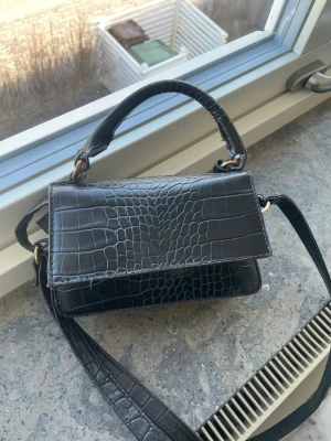 Svart handväska i krokopräglat skinn - Snygg svart handväska med krokodilpräglat mönster och både axelrem och handtag. Väskan har guldfärgade detaljer och en stilren, rektangulär form. Perfekt för dig som gillar trendiga accessoarer med lite edge.