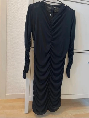 Svart långärmad omlottklänning - Snygg svart långärmad klänning med omlottdetalj och rynkade partier längs hela framsidan. Klänningen har v-ringning och är tillverkad i ett mjukt, stretchigt material som ger en smickrande passform.