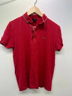Röd pikétröja från BOSS - Klassisk röd pikétröja från BOSS med korta ärmar och vit piping på kragen. Tröjan har knappslå framtill och diskret BOSS-logga broderad på bröstet. Perfekt till jeans eller shorts för en clean och stilren look.