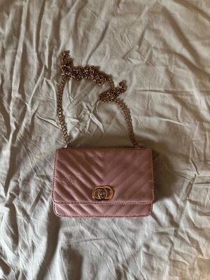 Rosa quiltad handväska med kedja - Snygg ljusrosa handväska med quiltat mönster och guldfärgad kedjerem. Väskan har ett dekorativt spänne framtill och flera fack inuti för kort och småsaker. Perfekt accessoar för att lyfta din outfit.