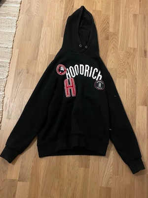 Svart hoodie från Hoodrich - Svart hoodie från Hoodrich med stor huva och broderade patches framtill. Stor vit text över bröstet, röd H och två runda märken. Klassisk känguruficka och ribbade muddar. Perfekt för en streetwear-look.