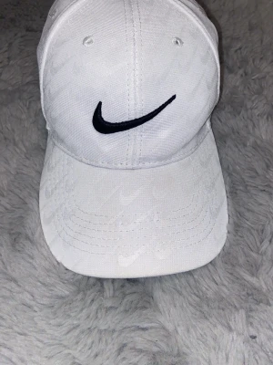 Vit keps från Nike med broderad logga - Snygg vit keps från Nike med svart broderad swoosh på framsidan. Kepsen har böjd skärm och diskret mönster av små swoosh-loggor över hela tyget. Tillverkad i bomullstyg och har ventilationshål för extra komfort.